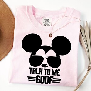Puede incluir: Una camiseta de color rosa claro con un gr&aacute;fico negro de una cabeza de rat&oacute;n con gafas de sol. Debajo del gr&aacute;fico, el texto dice TALK TO ME GOOF en letras may&uacute;sculas negras. La camiseta tiene una etiqueta de Comfort Colors en el cuello.