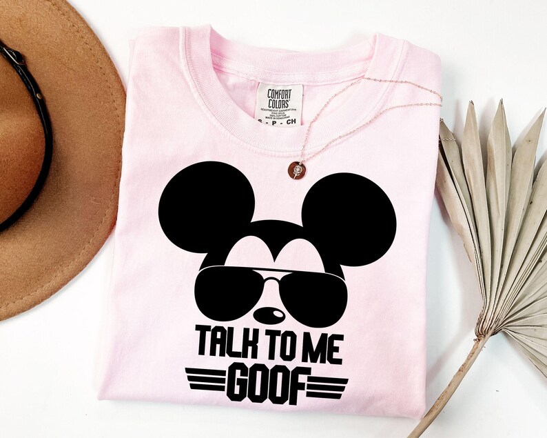 Puede incluir: Una camiseta de color rosa claro con un gr&aacute;fico negro de una cabeza de rat&oacute;n con gafas de sol. Debajo del gr&aacute;fico, el texto dice TALK TO ME GOOF en letras may&uacute;sculas negras. La camiseta tiene una etiqueta de Comfort Colors en el cuello.