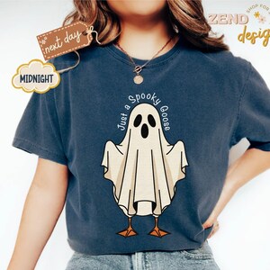May include: Navy blue t-shirt featuring a cartoon ghost with the text "Just a Spooky Goose". The ghost has black eyes and a beige body with orange feet. The shirt has a round neckline and short sleeves. The shirt also has the text "Midnight" and "next day" on a tag.