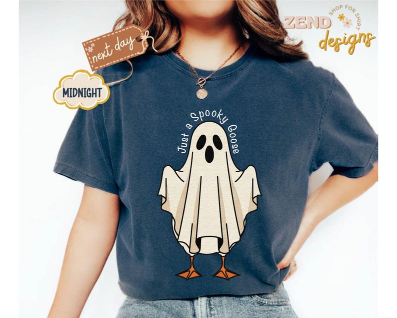 May include: Navy blue t-shirt featuring a cartoon ghost with the text "Just a Spooky Goose". The ghost has black eyes and a beige body with orange feet. The shirt has a round neckline and short sleeves. The shirt also has the text "Midnight" and "next day" on a tag.