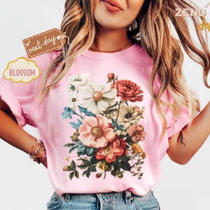 Puede incluir: Camiseta rosa claro con un dise&ntilde;o de ramo floral con flores blancas, rojas y melocot&oacute;n. La camiseta tiene mangas cortas y la palabra "Blossom" est&aacute; impresa en una etiqueta. Tambi&eacute;n se ve la marca "Zend Designs".