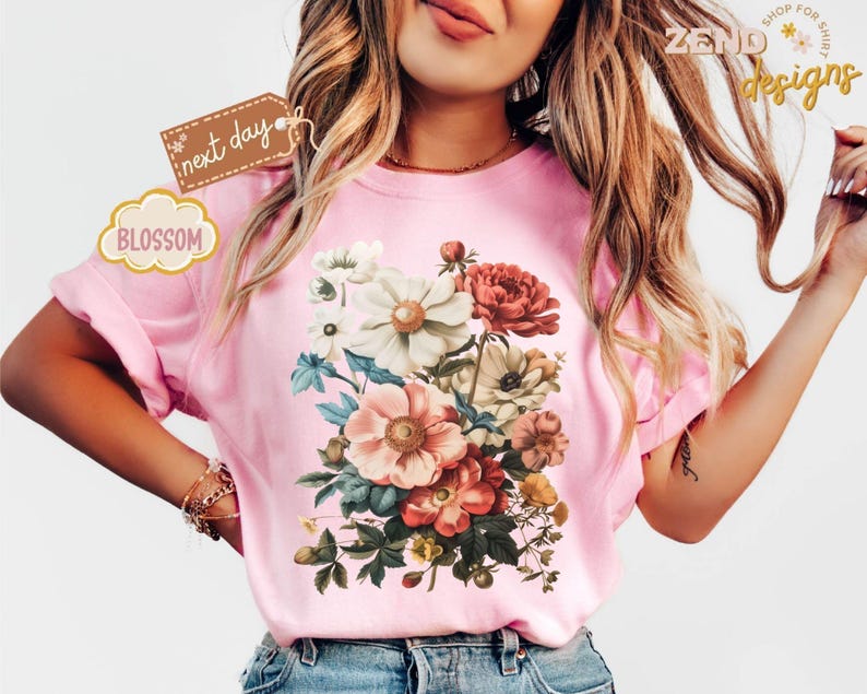 Puede incluir: Camiseta rosa claro con un dise&ntilde;o de ramo floral con flores blancas, rojas y melocot&oacute;n. La camiseta tiene mangas cortas y la palabra "Blossom" est&aacute; impresa en una etiqueta. Tambi&eacute;n se ve la marca "Zend Designs".