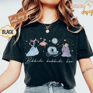 May include: Black t-shirt featuring a whimsical illustration of a fairytale scene with a dress, carriage, clock, and fairy godmother. The text "Bibbidi Bobbidi Boo" is written in a script font. The shirt has a vintage, worn look.