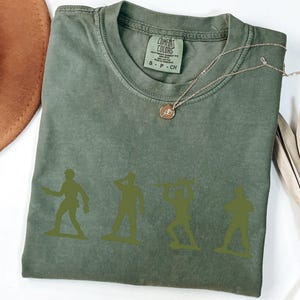 Puede incluir: Camiseta Comfort Colors verde oliva con cuatro siluetas de soldados de juguete verdes. La camiseta tiene cuello redondo y mangas cortas. Un collar dorado con un colgante est&aacute; drapeado sobre la camiseta. Un sombrero marr&oacute;n y una planta seca est&aacute;n en el fondo.