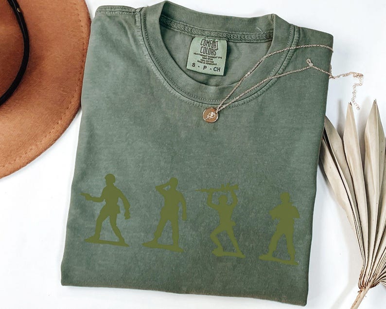 Puede incluir: Camiseta Comfort Colors verde oliva con cuatro siluetas de soldados de juguete verdes. La camiseta tiene cuello redondo y mangas cortas. Un collar dorado con un colgante est&aacute; drapeado sobre la camiseta. Un sombrero marr&oacute;n y una planta seca est&aacute;n en el fondo.