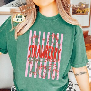 May include: A light green t-shirt with a pink and white striped design. The design features strawberries and the words "STRAWBERRY festival a berry good time" in red. The shirt has a relaxed fit.