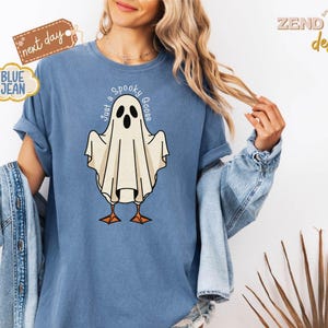 May include: Blue t-shirt with a ghost graphic that reads "Just a Spooky Goose." The ghost is white with black eyes and orange feet. The shirt has rolled-up sleeves and is worn with a denim jacket.
