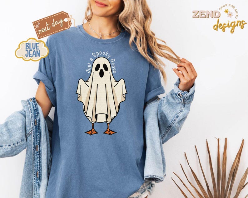 May include: Blue t-shirt with a ghost graphic that reads "Just a Spooky Goose." The ghost is white with black eyes and orange feet. The shirt has rolled-up sleeves and is worn with a denim jacket.