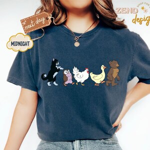 May include: Navy blue t-shirt featuring a whimsical illustration of a black and white cat, an owl, a white chicken, a yellow duck, and a brown bear walking together. The shirt has the text "ZEND designs" and "Midnight" on it.