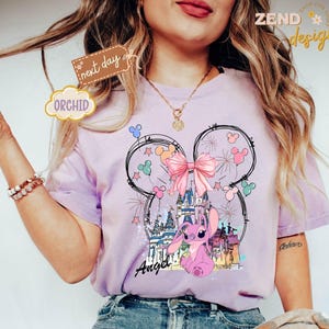 May include: A light purple t-shirt featuring a graphic of a pink cartoon character, a castle, and fireworks, with the word "Angel" in script. The design is set within a Mickey Mouse head outline, with a pink bow. The shirt has short sleeves.