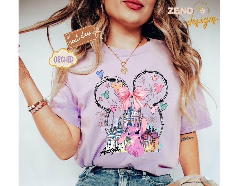May include: A light purple t-shirt featuring a graphic of a pink cartoon character, a castle, and fireworks, with the word "Angel" in script. The design is set within a Mickey Mouse head outline, with a pink bow. The shirt has short sleeves.