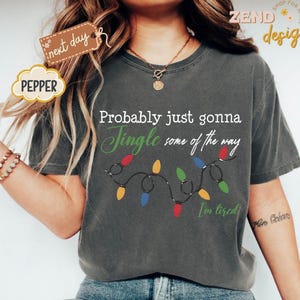Pode incluir: Camiseta cinza escuro com o texto "Probably just gonna Jingle some of the way" e uma sequ&ecirc;ncia de luzes de Natal. A camisa tamb&eacute;m tem o texto "I'm tired". A palavra "Pepper" est&aacute; em uma etiqueta.