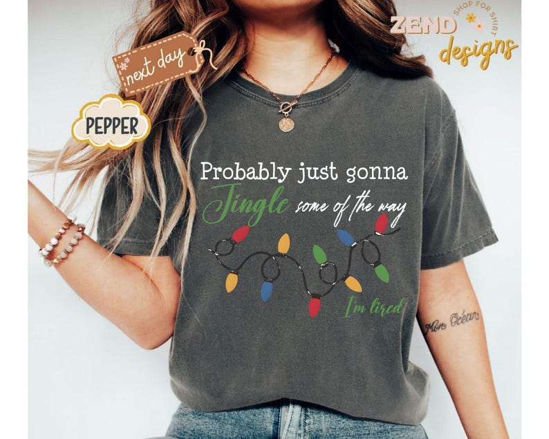 Pode incluir: Camiseta cinza escuro com o texto "Probably just gonna Jingle some of the way" e uma sequ&ecirc;ncia de luzes de Natal. A camisa tamb&eacute;m tem o texto "I'm tired". A palavra "Pepper" est&aacute; em uma etiqueta.