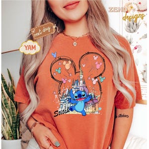 May include: Orange t-shirt featuring a graphic of Stitch from Lilo & Stitch. The design includes a castle, Mickey Mouse ears, and colorful balloons. The word "Stitch" is written in a script font below the character.