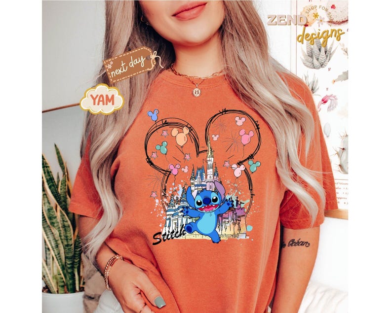May include: Orange t-shirt featuring a graphic of Stitch from Lilo & Stitch. The design includes a castle, Mickey Mouse ears, and colorful balloons. The word "Stitch" is written in a script font below the character.