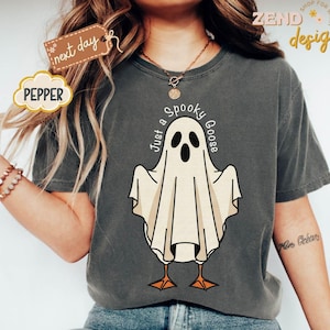 May include: Dark gray t-shirt with a cartoon ghost design. The ghost has a white sheet with a black face and orange feet. The text "Just a Spooky Goose" is above the ghost. The shirt has the word "Pepper" on it.