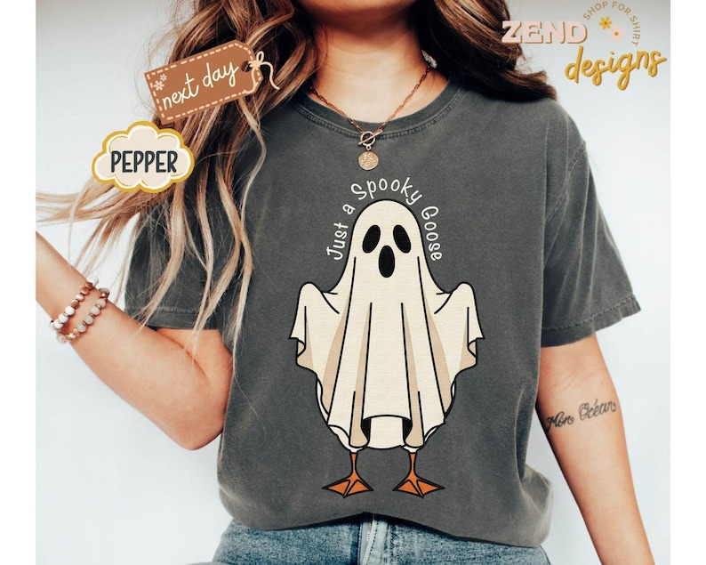 May include: Dark gray t-shirt with a cartoon ghost design. The ghost has a white sheet with a black face and orange feet. The text "Just a Spooky Goose" is above the ghost. The shirt has the word "Pepper" on it.