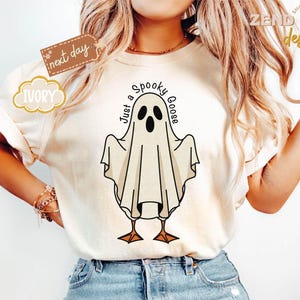 May include: Ivory-colored t-shirt featuring a whimsical ghost design with the text "Just a Spooky Goose." The ghost has black eyes and a beak, with a draped sheet-like body and orange feet. The shirt has short sleeves and a relaxed fit.