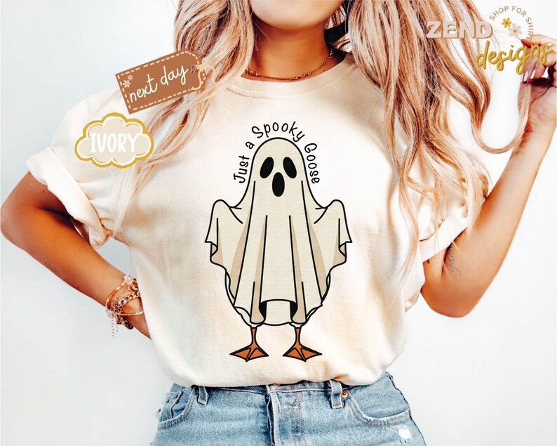 May include: Ivory-colored t-shirt featuring a whimsical ghost design with the text "Just a Spooky Goose." The ghost has black eyes and a beak, with a draped sheet-like body and orange feet. The shirt has short sleeves and a relaxed fit.
