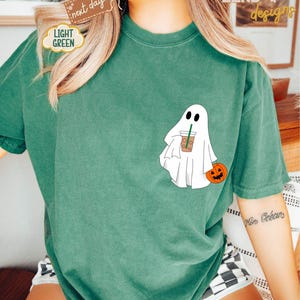 Pu&ograve; includere: Una maglietta verde con un fantasma bianco che tiene una tazza di caff&egrave; e una piccola zucca arancione. Il testo "ZEND designs" &egrave; nell'angolo in alto a destra dell'immagine.