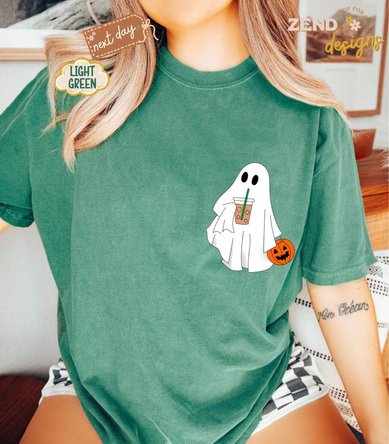 Pu&ograve; includere: Una maglietta verde con un fantasma bianco che tiene una tazza di caff&egrave; e una piccola zucca arancione. Il testo "ZEND designs" &egrave; nell'angolo in alto a destra dell'immagine.