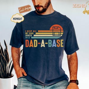 Könnte beinhalten: Marineblaues T-Shirt mit dem Text "I KEEP ALL MY DAD JOKES IN A DAD-A-BASE" in einem Retro-Design. Die Grafik enthält einen Sonnenuntergang und eine Server-Illustration. Ein bequemes, lässiges Kleidungsstück.