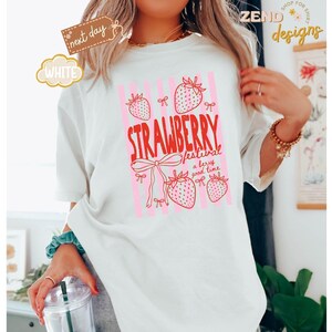 May include: White t-shirt with a pink and red strawberry design. The text reads "STRAWBERRY festival a berry good time". The shirt has a relaxed fit and short sleeves. The design includes strawberries and bows.