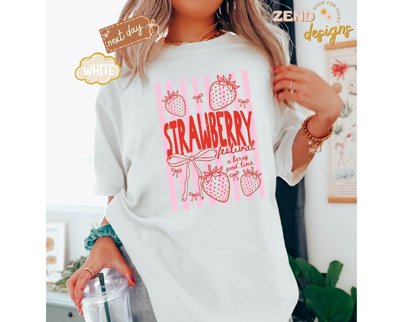 May include: White t-shirt with a pink and red strawberry design. The text reads "STRAWBERRY festival a berry good time". The shirt has a relaxed fit and short sleeves. The design includes strawberries and bows.