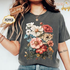 Puede incluir: Una camiseta gris oscuro con un dise&ntilde;o de ramo floral vibrante en tonos rosa, rojo, blanco y amarillo. La camiseta tiene el texto "Zend Designs" y "Pepper".