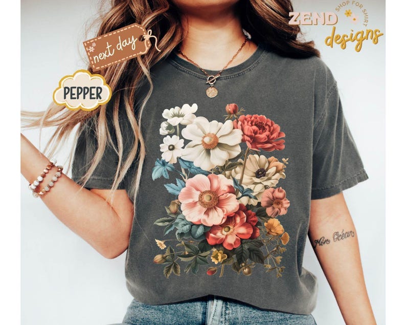 Puede incluir: Una camiseta gris oscuro con un dise&ntilde;o de ramo floral vibrante en tonos rosa, rojo, blanco y amarillo. La camiseta tiene el texto "Zend Designs" y "Pepper".