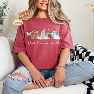 May include: Crimson-colored t-shirt featuring a Walt Disney World design. The graphic includes iconic landmarks and the text "Walt Disney World." The shirt is worn with ripped blue jeans. The shirt is from Zend Designs.