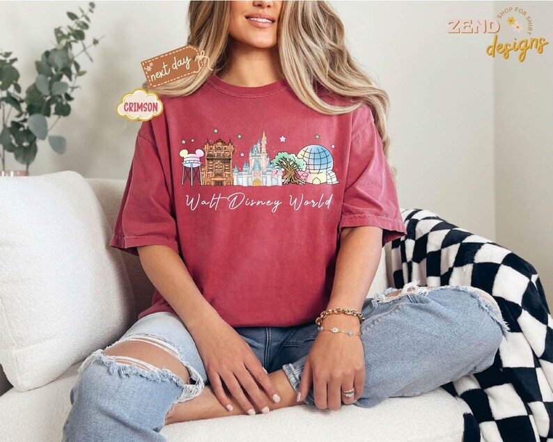 May include: Crimson-colored t-shirt featuring a Walt Disney World design. The graphic includes iconic landmarks and the text "Walt Disney World." The shirt is worn with ripped blue jeans. The shirt is from Zend Designs.