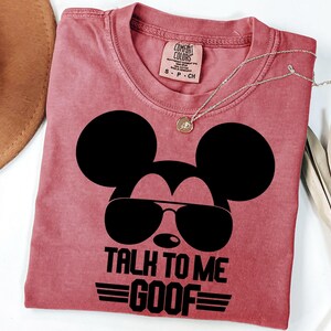 Puede incluir: Una camiseta de color rosa polvoriento con un gr&aacute;fico negro de la silueta de un rat&oacute;n con gafas de sol. El texto debajo dice TALK TO ME GOOF con l&iacute;neas horizontales a los lados. La camiseta tiene una etiqueta de marca Comfort Colors en la talla S.
