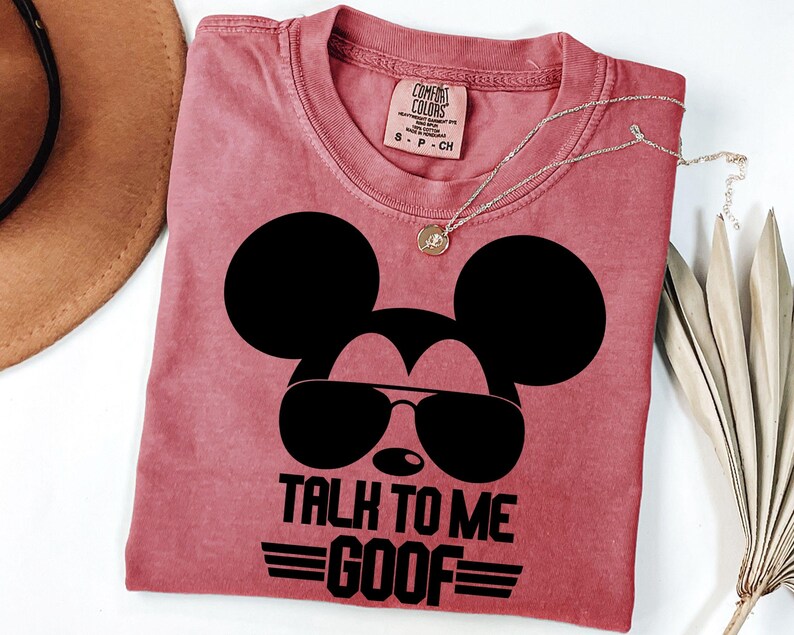 Puede incluir: Una camiseta de color rosa polvoriento con un gr&aacute;fico negro de la silueta de un rat&oacute;n con gafas de sol. El texto debajo dice TALK TO ME GOOF con l&iacute;neas horizontales a los lados. La camiseta tiene una etiqueta de marca Comfort Colors en la talla S.