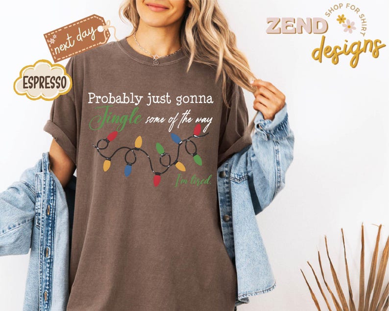 Pode incluir: Camiseta cor espresso com o texto "Probably just gonna Jingle some of the way" e uma sequ&ecirc;ncia de luzes de Natal coloridas. A camiseta tamb&eacute;m tem o texto "I'm tired". A camiseta faz parte da cole&ccedil;&atilde;o "Zend designs".
