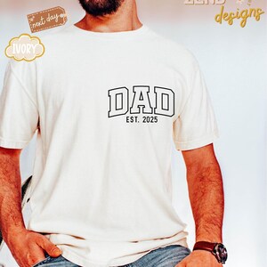 May include: White t-shirt with black text that reads "DAD EST. 2025".