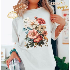 Puede incluir: Camiseta blanca con un dise&ntilde;o de ramo floral vintage en tonos rosa, rojo y crema. La camiseta tiene cuello redondo y mangas cortas. Las palabras "next day" y "white" est&aacute;n impresas en la camiseta.