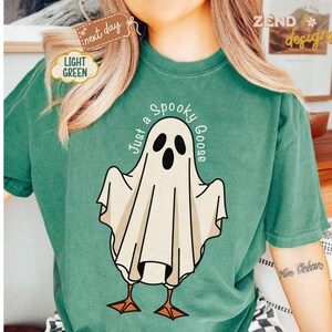 May include: A light green t-shirt featuring a cartoon ghost with the text "Just a Spooky Goose". The ghost has a white body, black eyes and mouth, and orange feet. The shirt has a relaxed fit and is made of a soft material.
