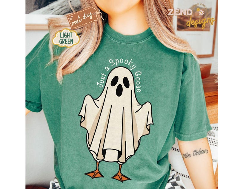 May include: A light green t-shirt featuring a cartoon ghost with the text "Just a Spooky Goose". The ghost has a white body, black eyes and mouth, and orange feet. The shirt has a relaxed fit and is made of a soft material.