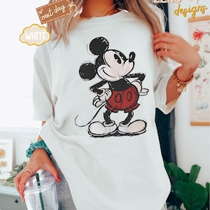 May include: White t-shirt with a vintage-style illustration of Mickey Mouse in black and red. The illustration shows Mickey Mouse standing with his right hand on his hip and his left hand holding his tail.