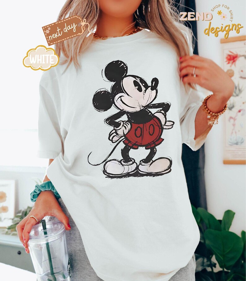 May include: White t-shirt with a vintage-style illustration of Mickey Mouse in black and red. The illustration shows Mickey Mouse standing with his right hand on his hip and his left hand holding his tail.