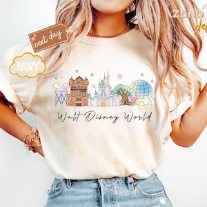 May include: Ivory-colored t-shirt featuring a colorful illustration of Walt Disney World landmarks, including the Tower of Terror, Cinderella Castle, and Spaceship Earth. The words "Walt Disney World" are written in a script font below the graphic. The shirt has rolled-up sleeves.