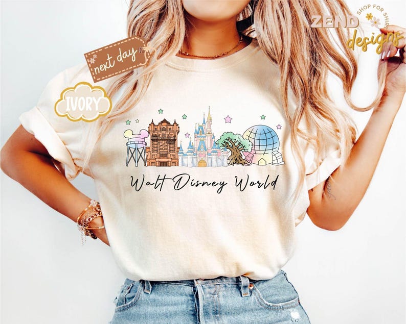 May include: Ivory-colored t-shirt featuring a colorful illustration of Walt Disney World landmarks, including the Tower of Terror, Cinderella Castle, and Spaceship Earth. The words "Walt Disney World" are written in a script font below the graphic. The shirt has rolled-up sleeves.