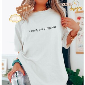 May include: White t-shirt with the text "i can't, i'm pregnant" in black. The shirt has short sleeves and a relaxed fit. The image also includes a clear plastic cup with a straw and a teal scrunchie.