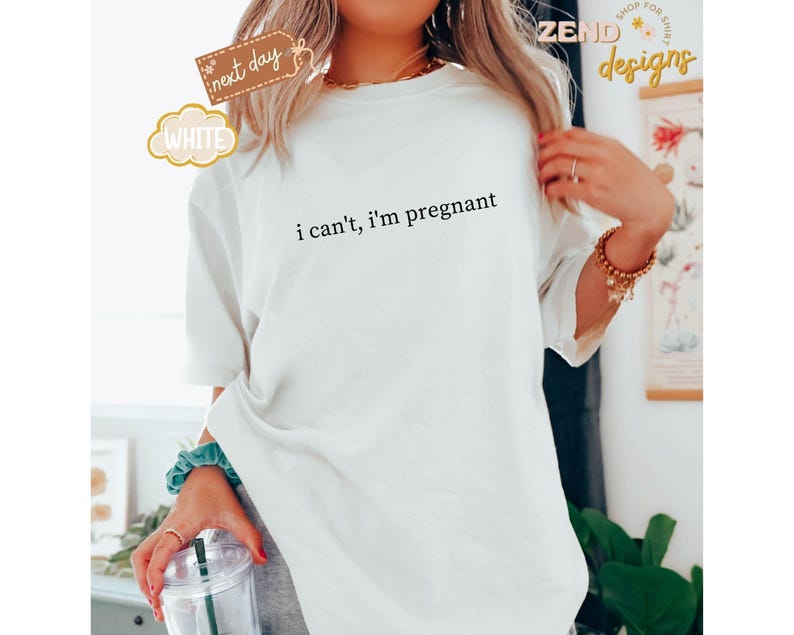 May include: White t-shirt with the text "i can't, i'm pregnant" in black. The shirt has short sleeves and a relaxed fit. The image also includes a clear plastic cup with a straw and a teal scrunchie.