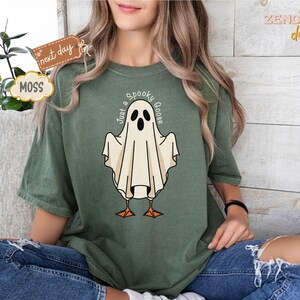May include: Moss green t-shirt featuring a cartoon ghost with the text "Just a Spooky Goose". The ghost has black eyes and a beak, with orange feet. The shirt is a relaxed fit and is worn with blue jeans.