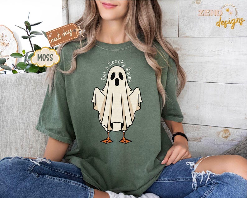 May include: Moss green t-shirt featuring a cartoon ghost with the text "Just a Spooky Goose". The ghost has black eyes and a beak, with orange feet. The shirt is a relaxed fit and is worn with blue jeans.