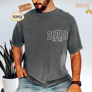 May include: A charcoal gray t-shirt with a white graphic that says "DAD EST. 2025".