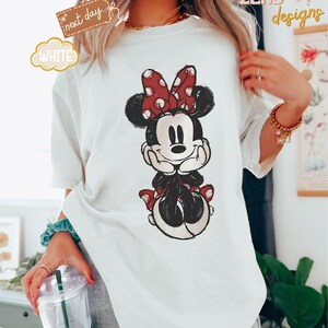 May include: White t-shirt with a cartoon image of Minnie Mouse wearing a red bow with white polka dots. Minnie Mouse is sitting with her hands on her chin and has a happy expression on her face.