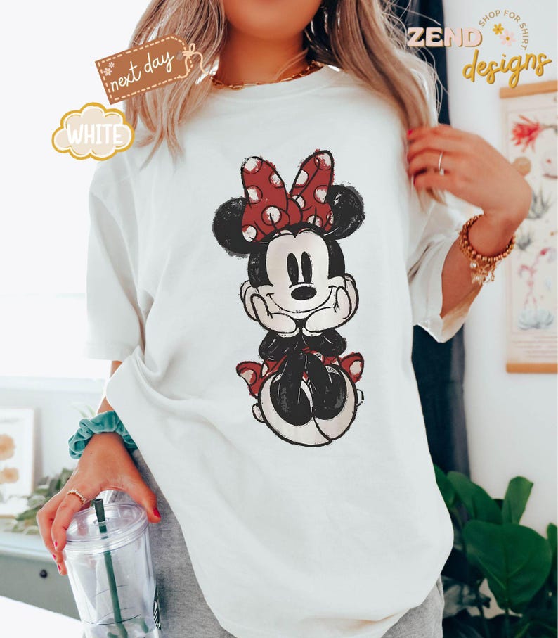 May include: White t-shirt with a cartoon image of Minnie Mouse wearing a red bow with white polka dots. Minnie Mouse is sitting with her hands on her chin and has a happy expression on her face.